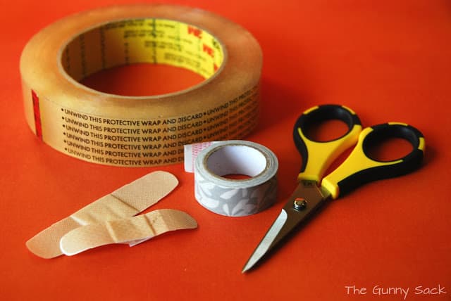 Double-sided tape, a pair of scissors, a roll of Washi tape and two bandages on a countertop.