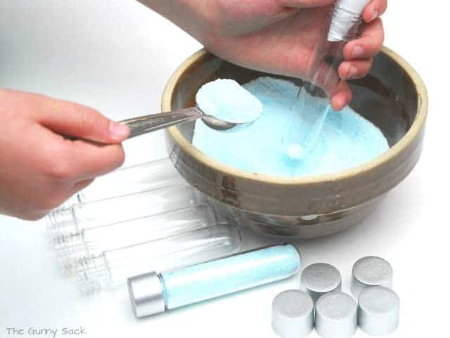 hands using a spoon and a funnel to add bath salts to a clear plastic test tube.